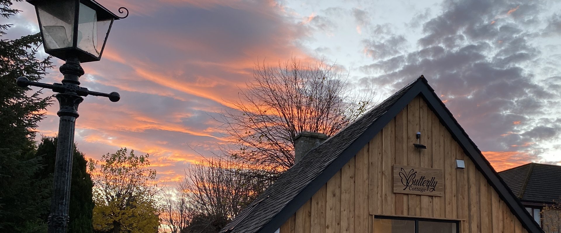 Butterfly Cottage bij zonsondergang, de gevel met lariksbekleding en het bord Butterfly Cottage onder een oranje gestreepte lucht